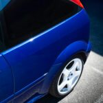 Close-up of a blue sports car with white rims shining in sunlight, outdoors.